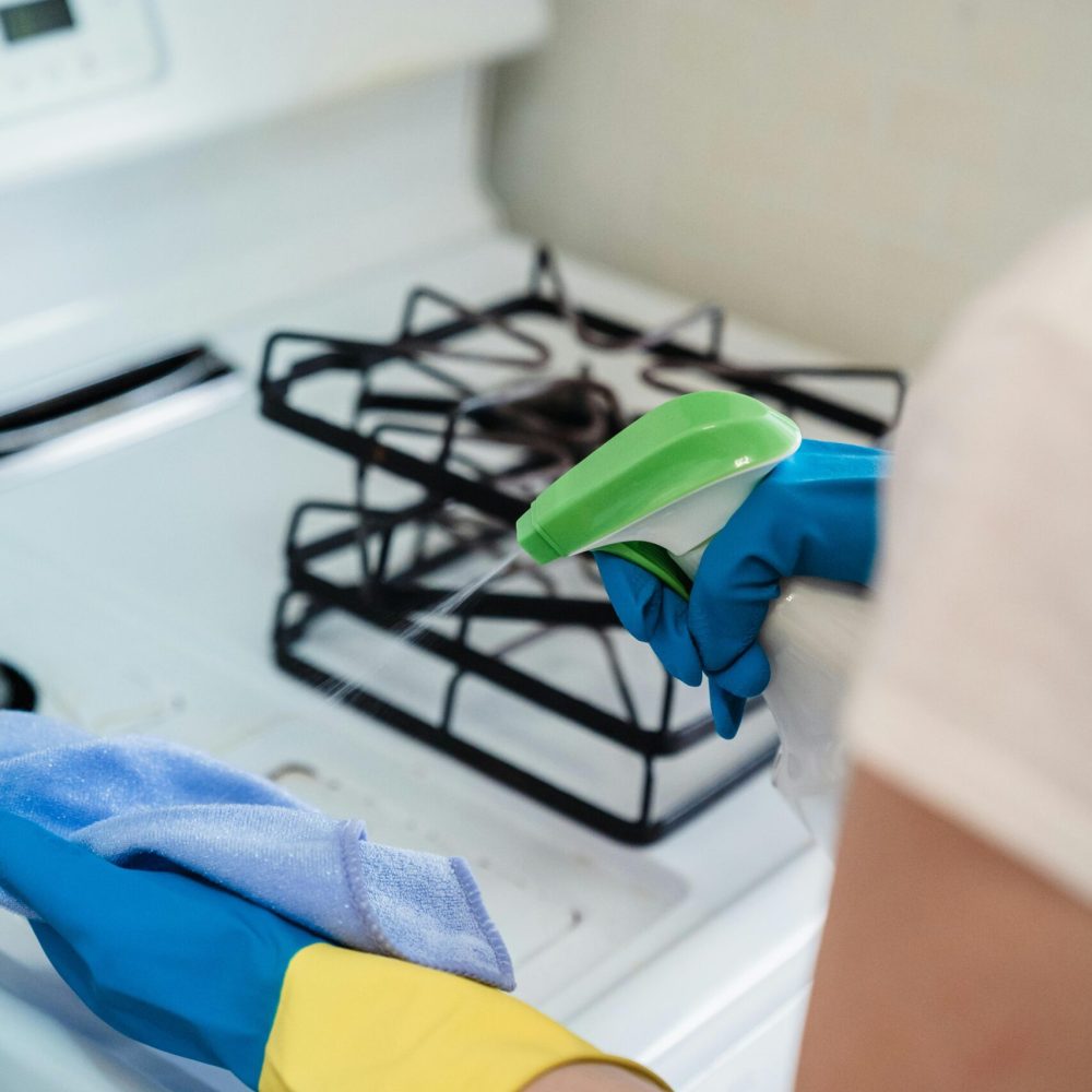 A person wearing gloves uses a spray bottle and cloth to clean a kitchen stove.
